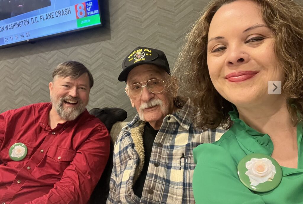 A group photo of three individuals smiling, one wearing a 'Vietnam Era Veteran' cap and another with a flower brooch on their shirt.
